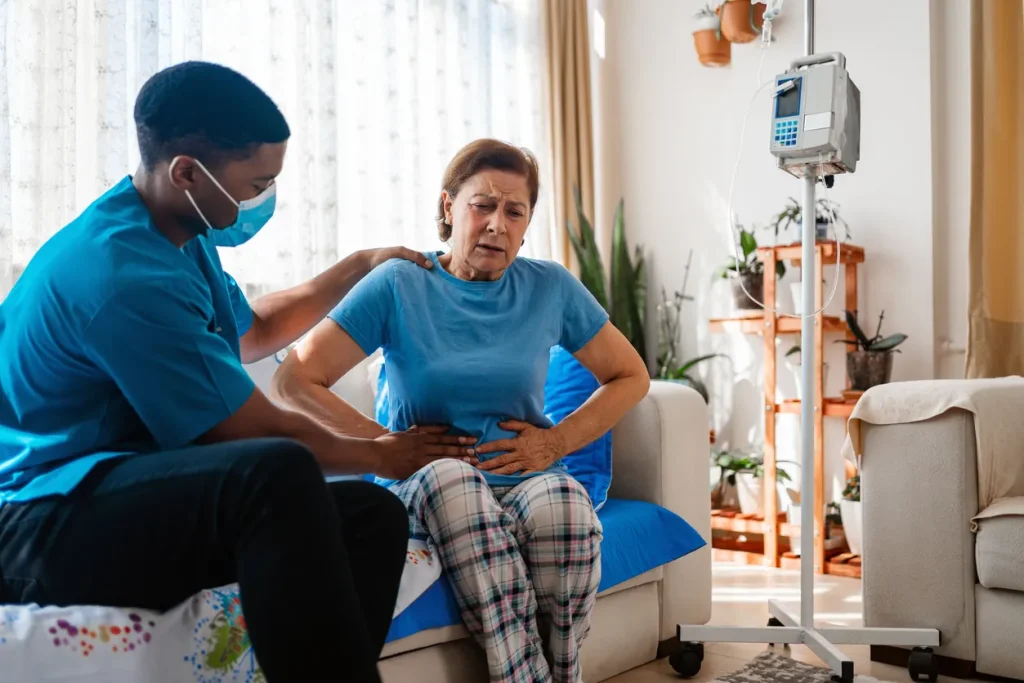 Image of an older patient sitting on a bed, clutching her abdominal region, being assisted by a nurse, illustrating pancreatic cancer, diarrhea symptoms, exocrine pancreatic insufficiency, cancer-related digestive issues, and pancreatic enzyme deficiency.