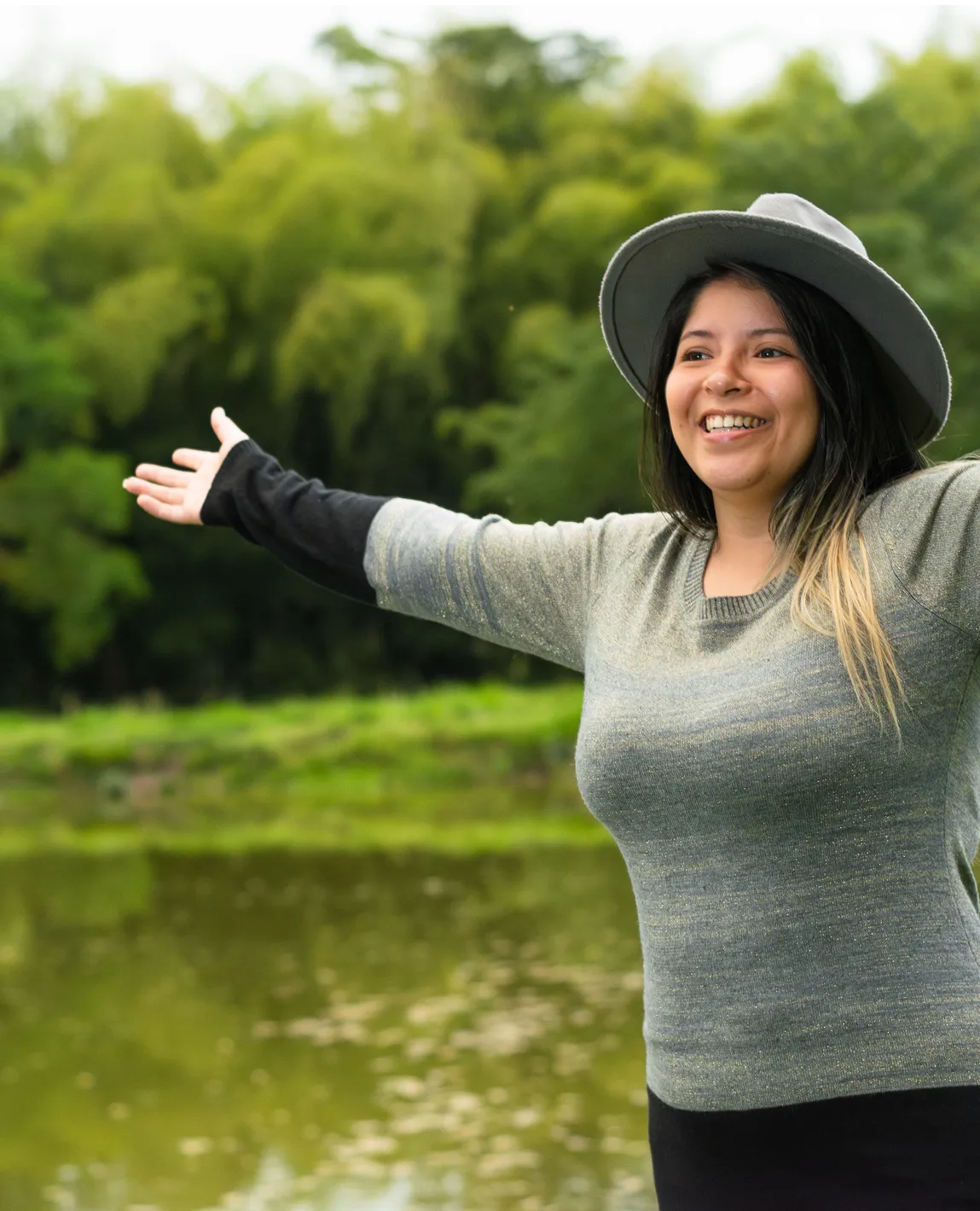 A smiling woman stands in front of a pond with her arms outstretched.