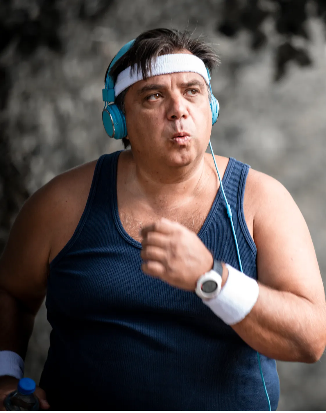 A large man wearing a tanktop, sweatbands on his wrists and head, and headphones going for a run.