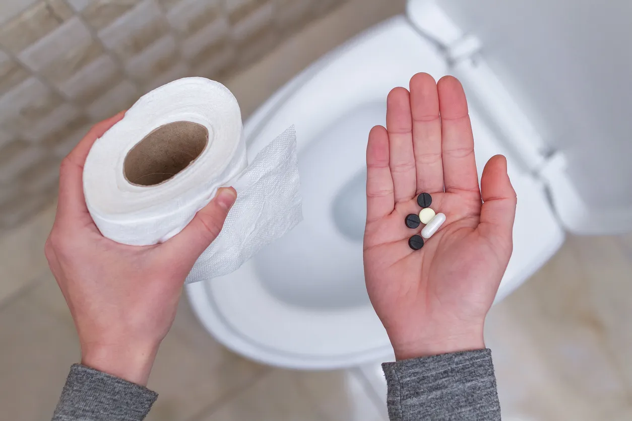 A person standing next to a toilet, holding toilet paper in one hand and pills in the other, representing various diarrhea treatment options.