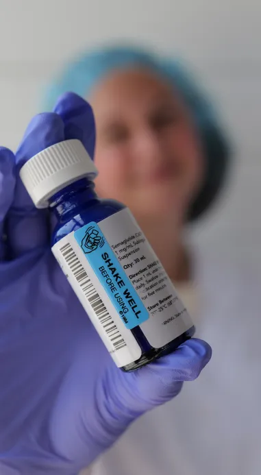 A woman in a lab coat, and gloves holds a bottle of medicine.