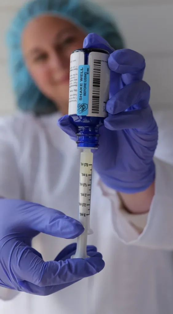 A pharmacy technician, wears a lab coat, gloves, and hair covering, while demonstrating how to put medication into the syringe.